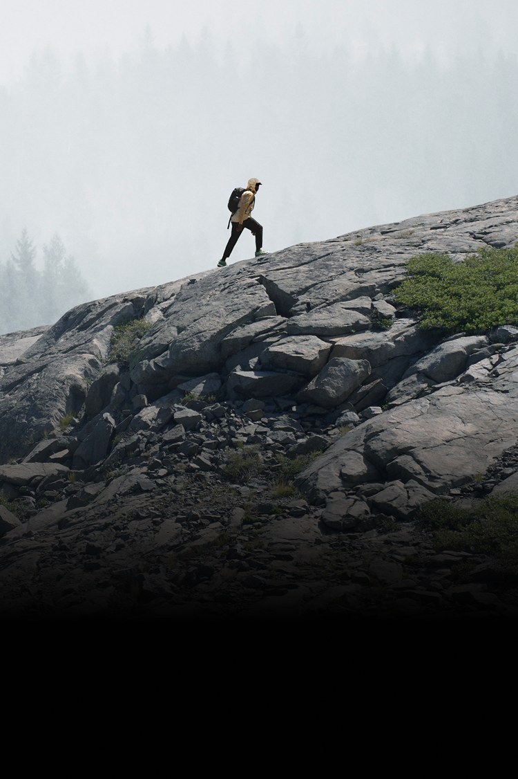 A person in a rain jacket from The North Face hikes up a rocky ridge. In the background, gray mist covers the trees.