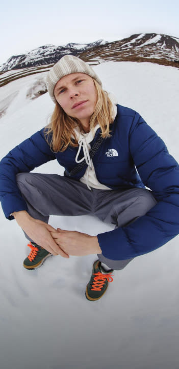 A guy in a white hoodie/tan beanie/navy jacket sitting in the snow. He’s looking away from the camera.