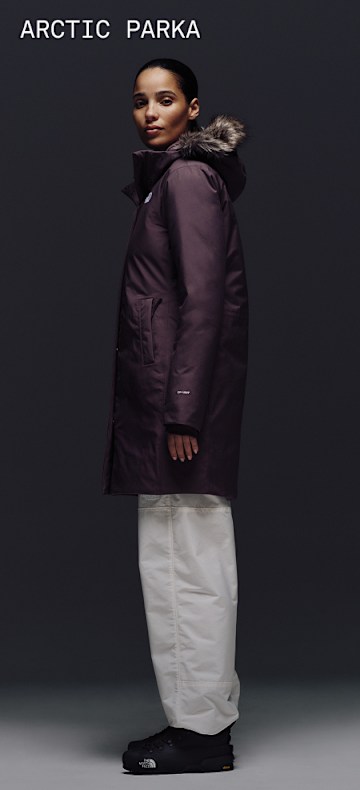 Female model wearing The North Face Arctic Parka in Tawny Quartz with fur-trimmed hood, shown against a dark backdrop. 