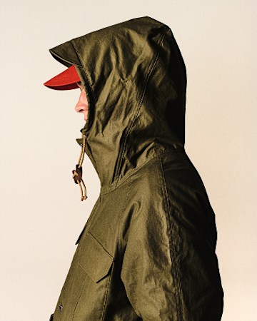 Cream background with man facing the right showing his nose / profile, wearing the army-green parka with the hood pulled up and the red hat.