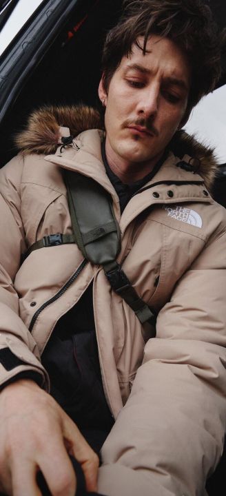 A guy in a white hoodie/tan beanie/navy jacket sitting in the snow. He’s looking away from the camera.