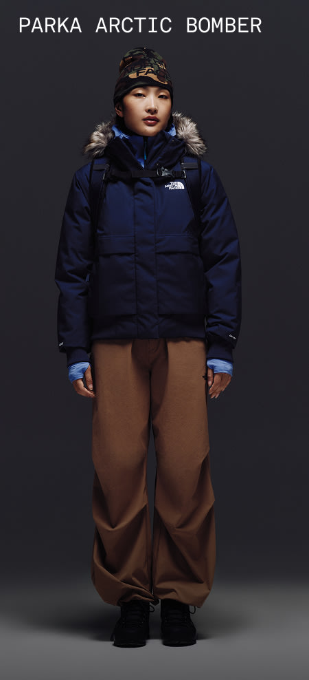 Une mannequin portant le blouson Arctic The North Face dans la couleur sommet marine avec un capuchon bordé de fourrure et un pantalon de neige brun.