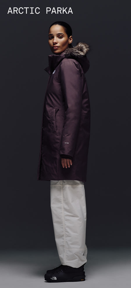 Female model wearing The North Face Arctic Parka in Tawny Quartz with fur-trimmed hood, shown against a dark backdrop. 