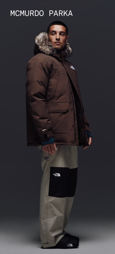 Male model wearing The North Face McMurdo Parka in Smokey Brown with fur-trimmed hood.