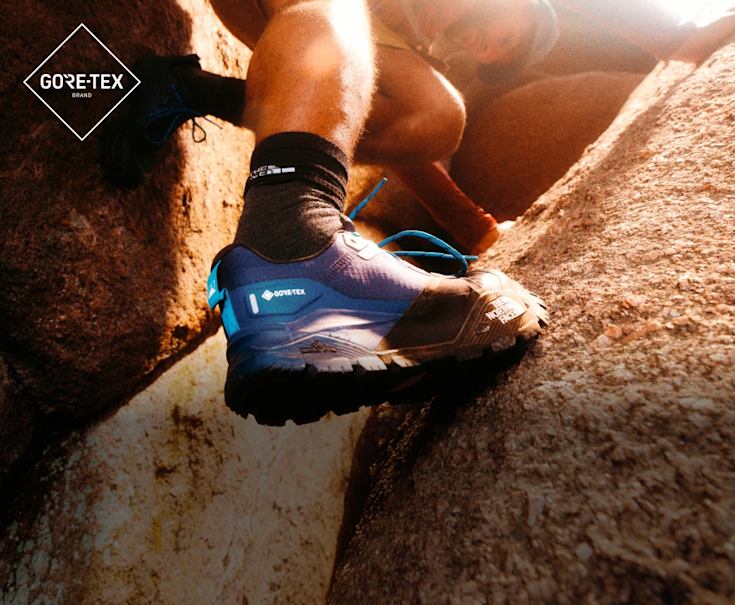 A man wearing low-rise, blue hiking shoes with black socks. He’s scaling red rocks. 
