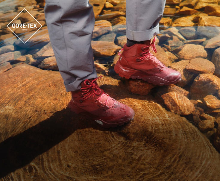 A woman wearing low-rise, tan hiking shoes with black and gray socks. She’s scaling red rocks. 