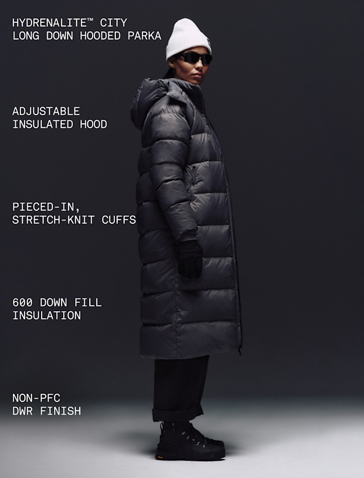 A female wearing The North Face Hydrenalite City Long Down Hooded Parka in Tawny Quartz and a black knit beanie looks down against a neutral background. 