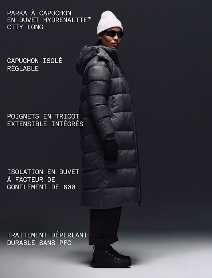 A female wearing The North Face Hydrenalite City Long Down Hooded Parka in Tawny Quartz and a black knit beanie looks down against a neutral background. 
