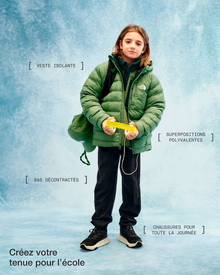 Un enfant vêtu d’un manteau vert, d’un pantalon noir et de chaussures noires, tenant des écouteurs et regardant l’appareil photo, avec un léger sourire.