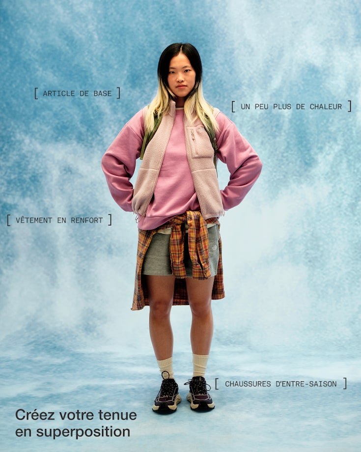 Personne vêtue d’un chandail molletonné rose, d’une veste beige, d’un short et de chaussures noires, avec une chemise à carreaux nouée autour de la taille, les mains sur les hanches, regardant droit vers la caméra.