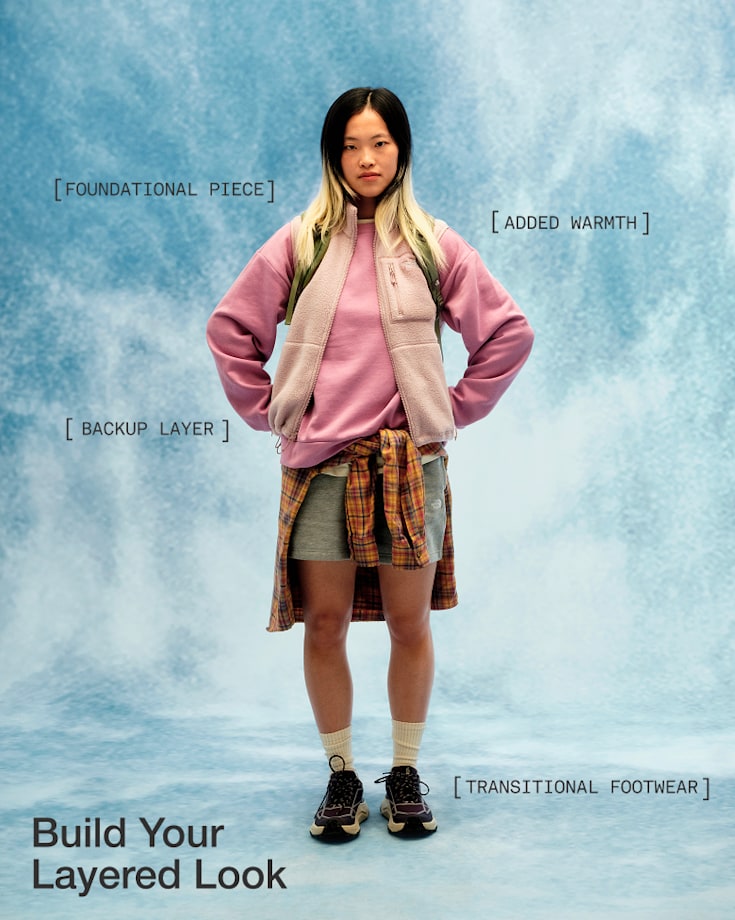 Person in a pink sweatshirt, beige vest, shorts and black shoes with a plaid shirt tied around the waist, hands on hips and looking directly at the camera.