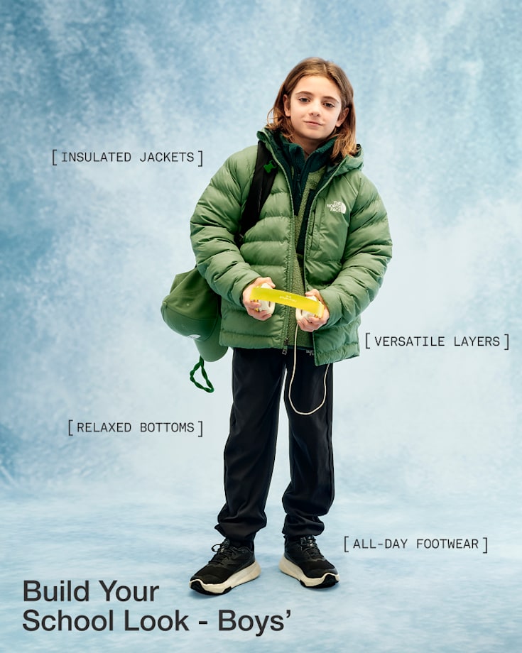 A child in a green jacket, black pants and black shoes, holding a pair of headphones and looking at the camera, slightly smiling.