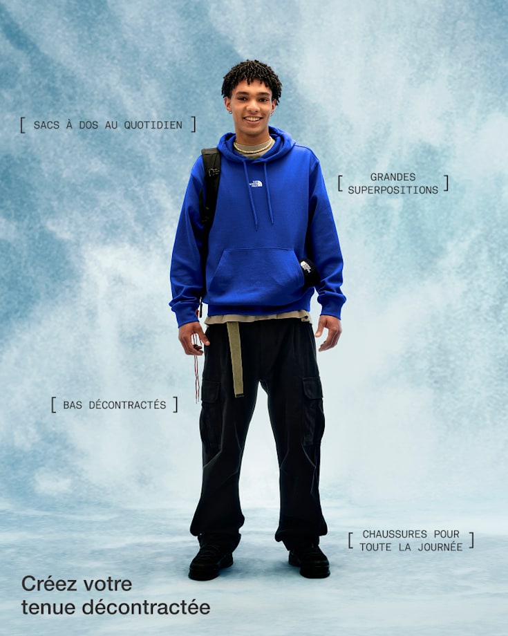 Une personne vêtue d’un chandail à capuchon bleu, d’un pantalon noir et de chaussures noires, qui sourit et regarde l’appareil photo.