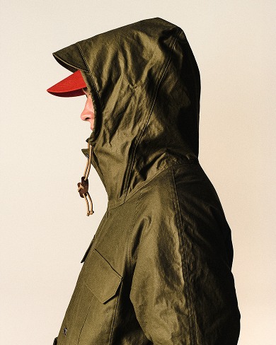 Cream background with man facing the right showing his nose / profile, wearing the army-green parka with the hood pulled up and the red hat.