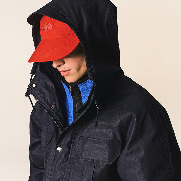 Cream background with man facing the right showing his nose / profile, wearing the army-green parka with the hood pulled up and the red hat.