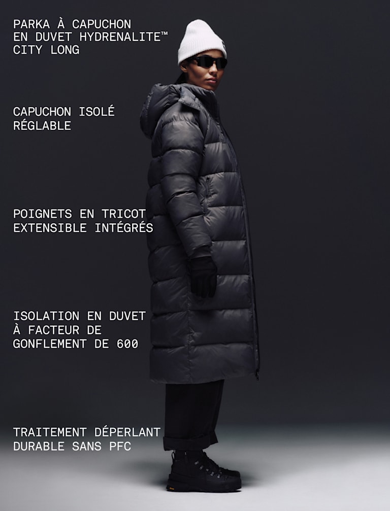 A female wearing The North Face Hydrenalite City Long Down Hooded Parka in Tawny Quartz and a black knit beanie looks down against a neutral background.