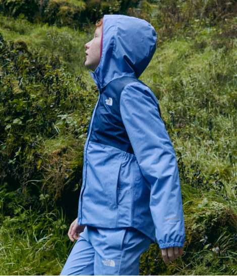 The backside of a kid in a two-toned blue rain jacket climbing down rocks to a rocky beach. 
