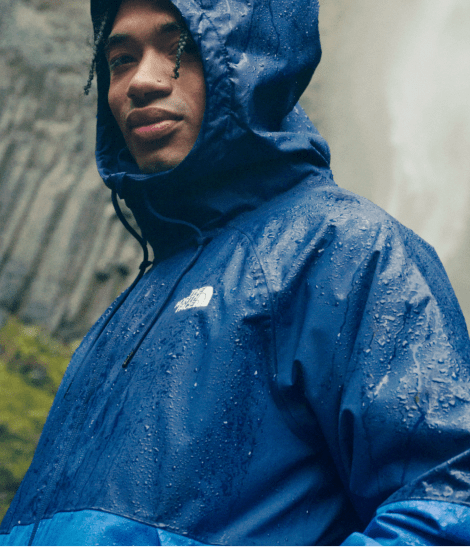A man in a two-toned blue rain jacket with the hood up and waterfall behind him.