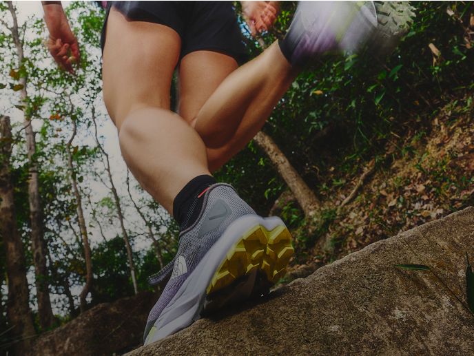 A photo of a runner from beneath them. They’re running through a green forest and is wearing light purple shoes, black socks and black running shorts.