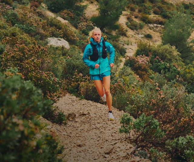 A woman in an all-aqua running outfit and black running vest running through green shrubbery.