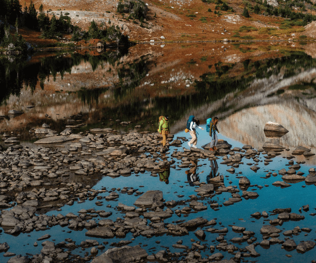 Three hikers wearing gear from The North Face pick their way across a rocky body of water.