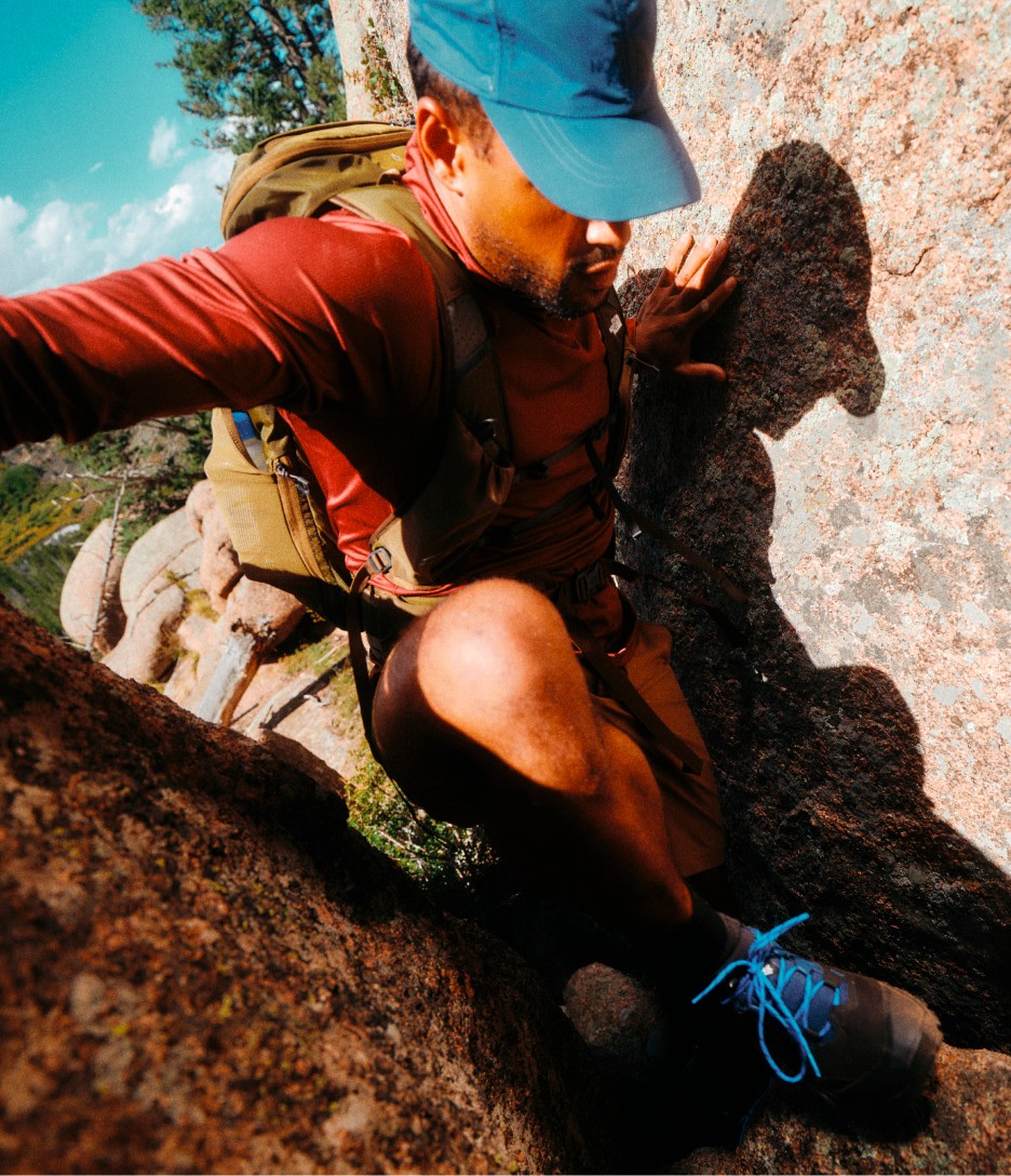 A man in a blue hat and red long-sleeved shirt stepping up onto a rock. 