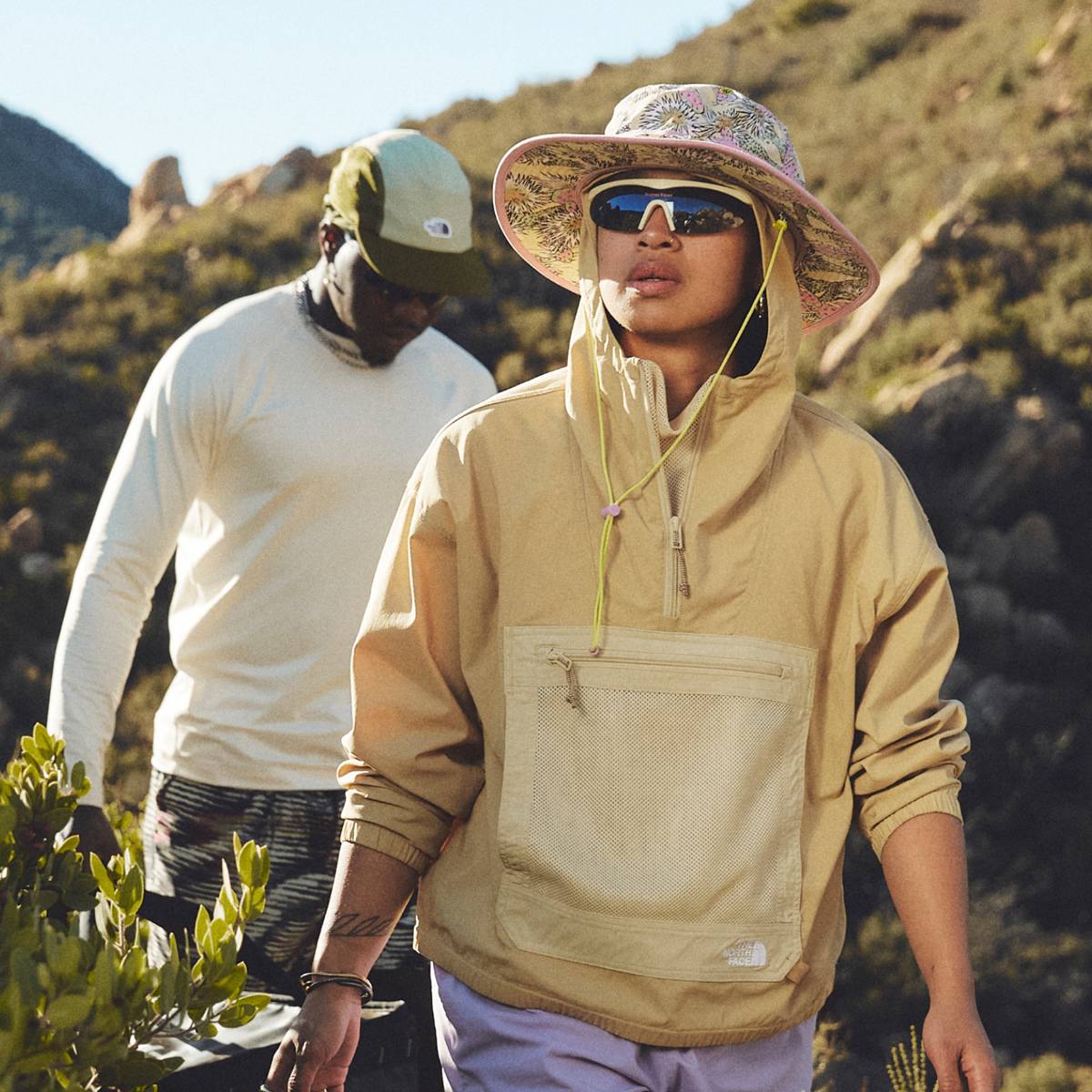 Two hikers walk through a sunny mountain landscape wearing hats from The North Face.