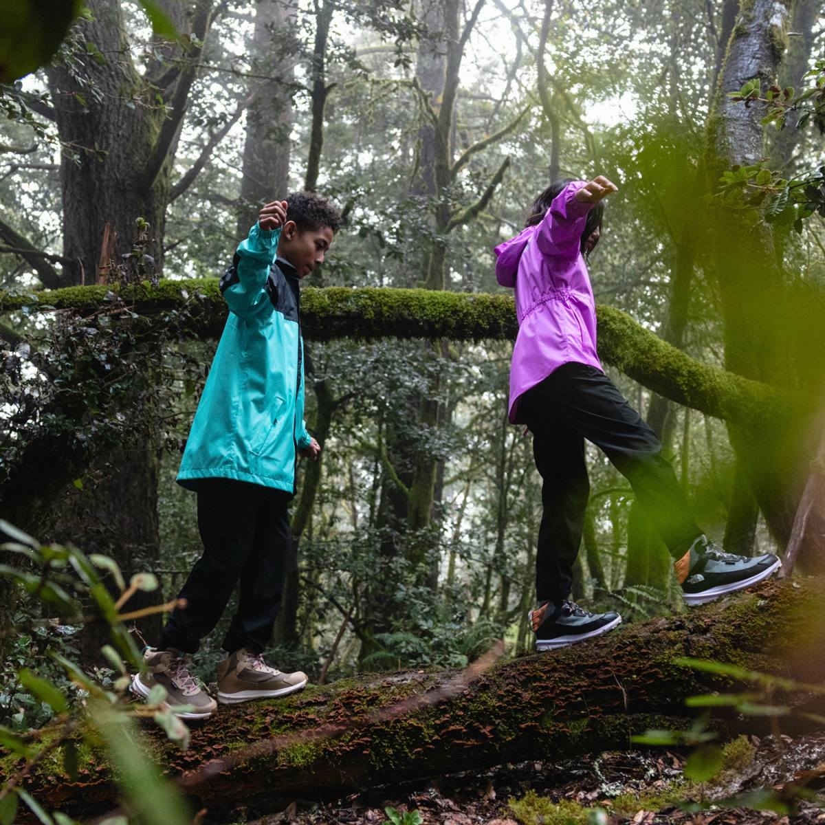 Kids wearing hiking boots from The North Face walk along a mossy log in a forest. 
