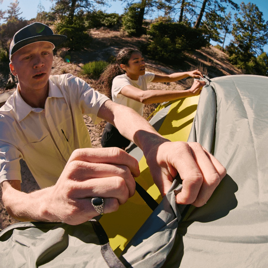 Two campers set up a tent at a sunny campground. 