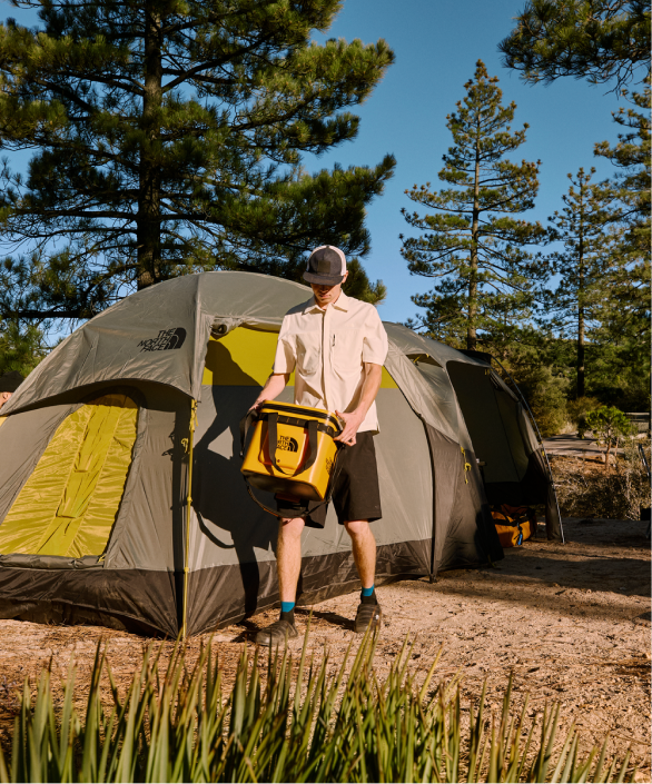 A man carries a cooler by The North Face in front of a camping tent.