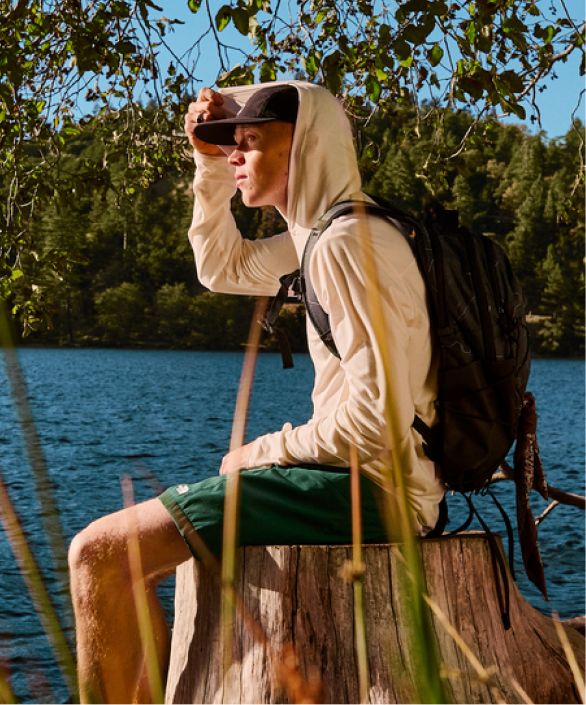 A man sits on a the side of a body of water using binoculars. 