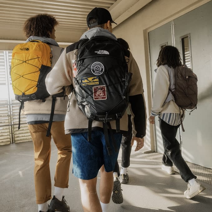 Kids walking past school doors. The one on the left has a yellow backpack, the one in the center has a backpack with patches and the one on the right has a brown backpack. 