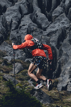 The backside of a man running up a mountain in a red jacket, floral vest and running shorts, and hiking polls.