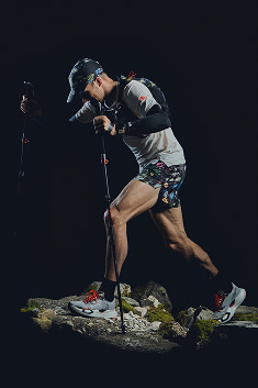 A man running up a mountain in a floral vest and running shorts, and hiking polls.