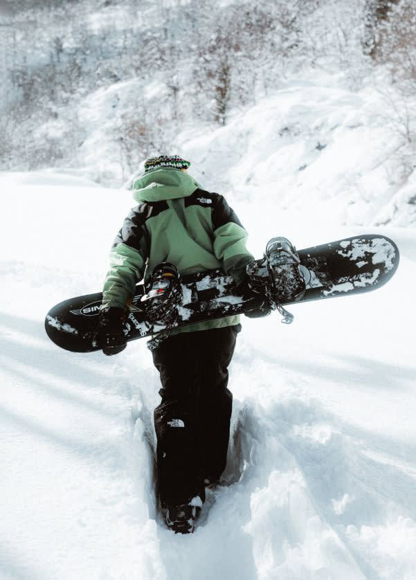 Image of a person wearing The North Face hiking up a snow covered mountain while carrying a snowboard.