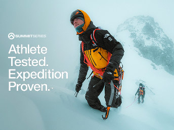 A man in a yellow and black jacket trudging up a snow-covered hill with rope sashed across him. Someone is following behind.