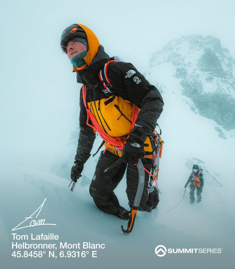 A man in a yellow and black jacket trudging up a snow-covered hill with rope sashed across him. Someone is following behind.
