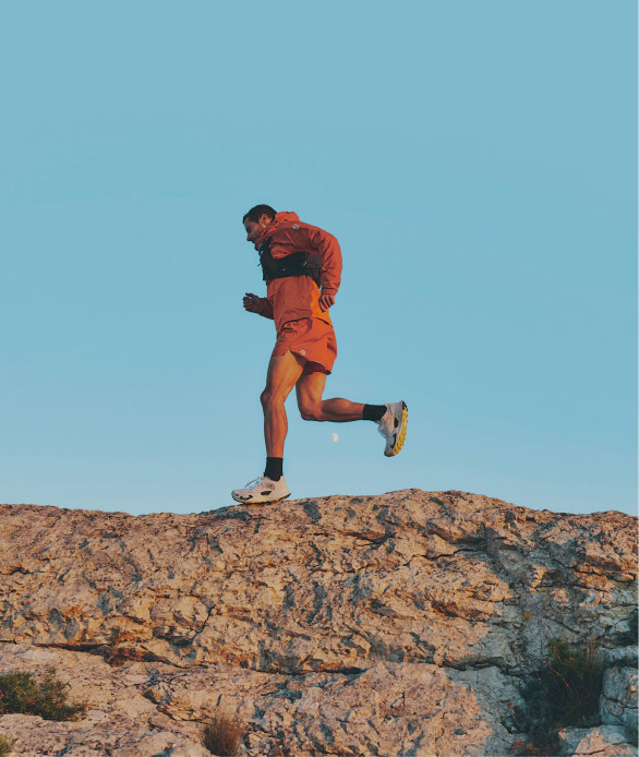 A woman with black hair in a ponytail and white half zip fleece walking through greenery.