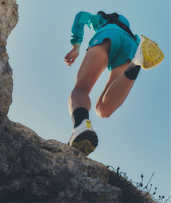 A woman with black hair in a ponytail and white half zip fleece walking through greenery.