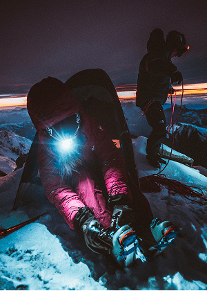 A woman in a pink jacket and pink flat bill hat sticking out from under her hood. She has sunglasses on and skis on her back. One hand is holding a hiking poll. 