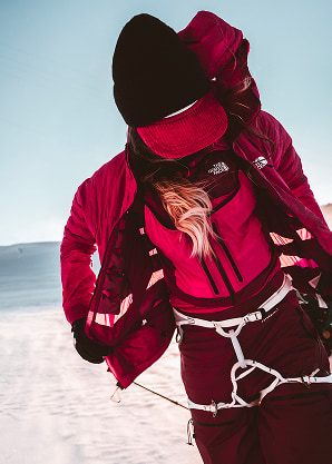 A woman in a pink kit standing with her jacket open revealing a pink vest. She’s wearing a climbing harness.