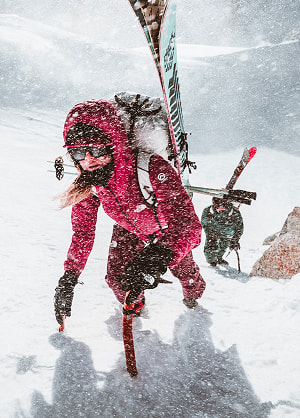 A woman in an all pink kit with sunglasses and skis on her back hiking up a snowy mountain side. It’s actively snowing.
