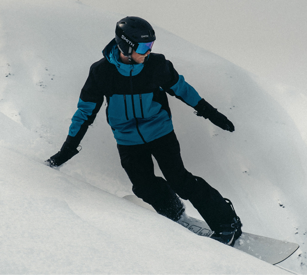 A snowboarder wearing The North Face Men’s Chakal Jacket in Dusk Blue/TNF Black carves down a snowy mountain slope, surrounded by pristine powder and rugged alpine peaks.