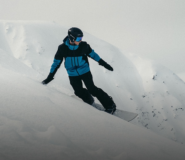Un planchiste portant le manteau Chakal de The North Face pour hommes dans la couleur bleu crépuscule/noir TNF dévale une pente raide en glissant sur la neige profonde. 