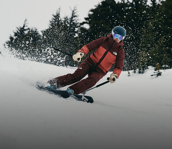 Une skieuse portant le manteau THERMOBALL™ Snow Triclimate® de The North Face pour femmes dans la couleur sumac/poussière de Mars fend la poudreuse et projette de la neige tout en dévalant la montagne. 