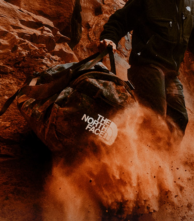 Person dragging a dirt-covered Base Camp Duffel across red rock terrain, sending dust into the air. 