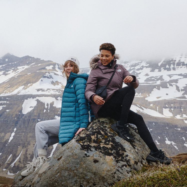 2 women sitting in a tundra with a snow capped mountain behind them