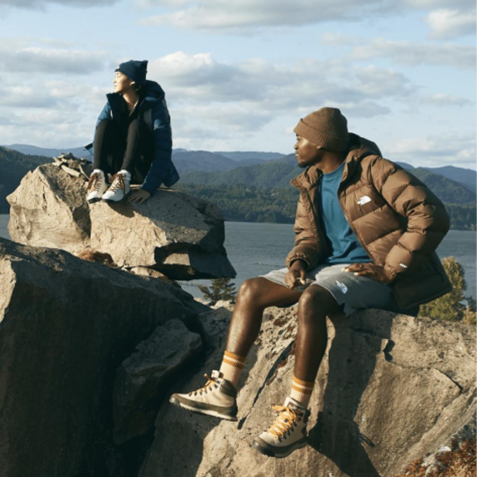 man and woman sitting on top of a rock next to lake and mountains