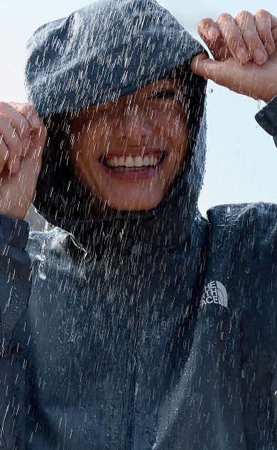 Une femme sourit en tenant sa capuche sous la pluie.