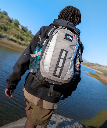 Person walking along a river wearing a black jacket and gray backpack.